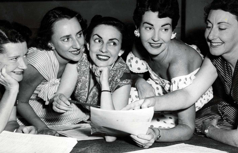 Five women leaning against a desk looking at a sheet of paper, they are all smiling.